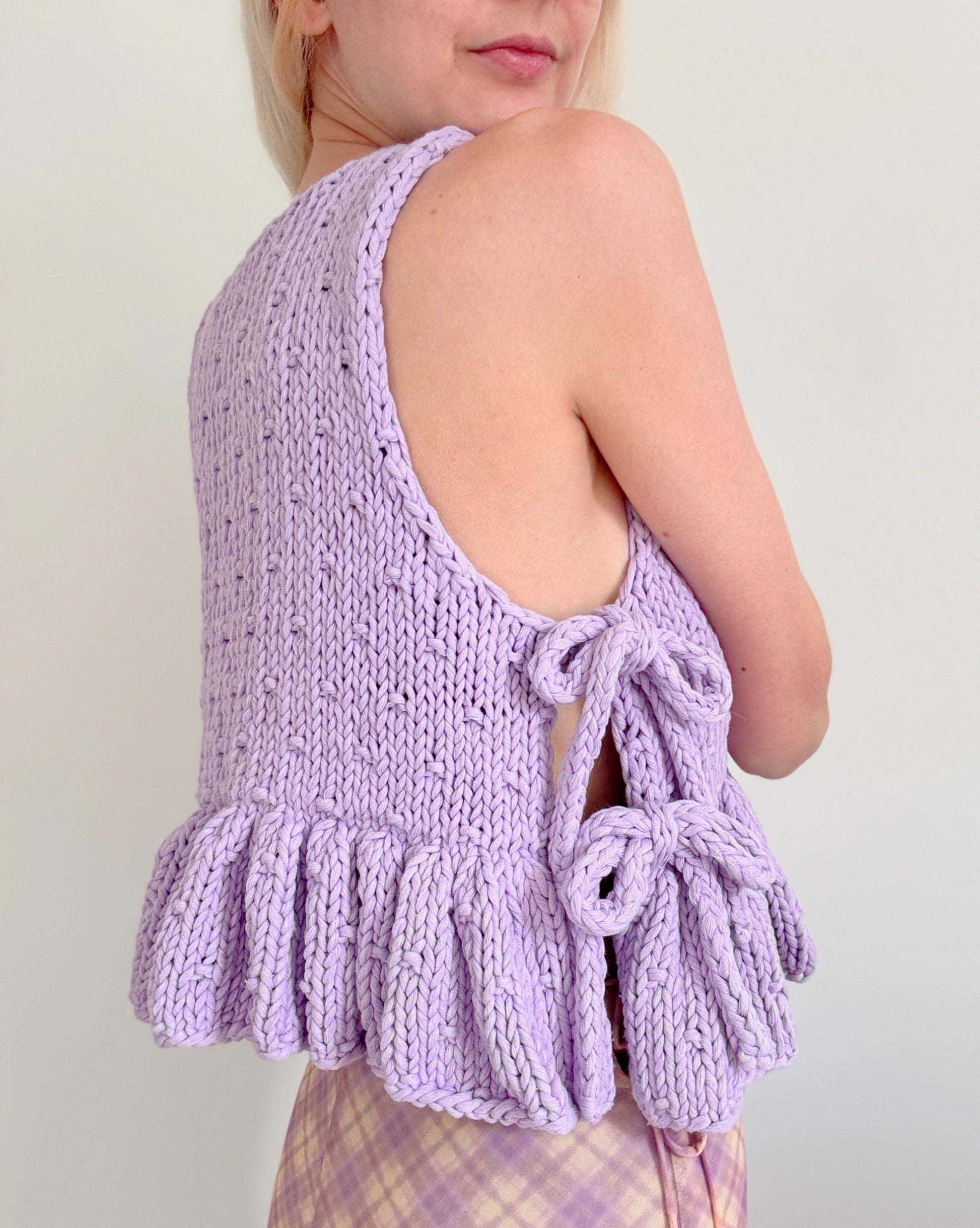 Person wearing a purple knitted sleeveless top with ruffled details on a plain background